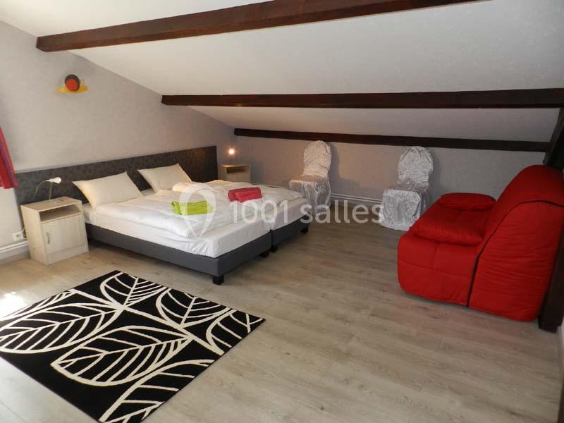 Chambre mansardée avec lit double, fauteuil rouge, deux chaises blanches, tapis à motifs et lumière naturelle.