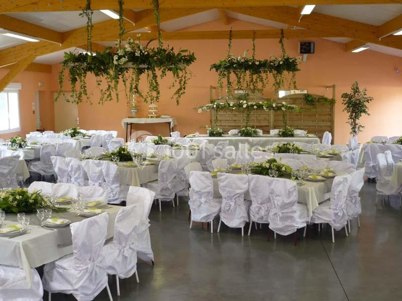 Salle de réception décorée avec des tables dressées, des chaises couvertes de housses blanches et des suspensions végétales.