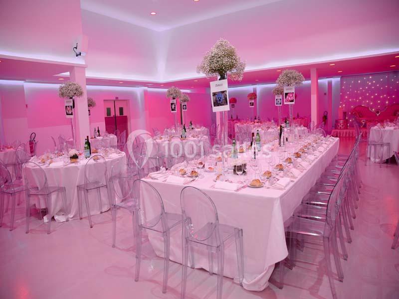 Salle de réception décorée en rose avec des tables rondes dressées, des chaises transparentes et des centres de table…