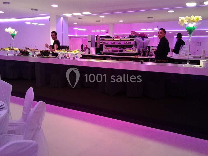 Salle de réception moderne avec un long bar éclairé en violet, des serveurs et des décorations florales sur les tables.