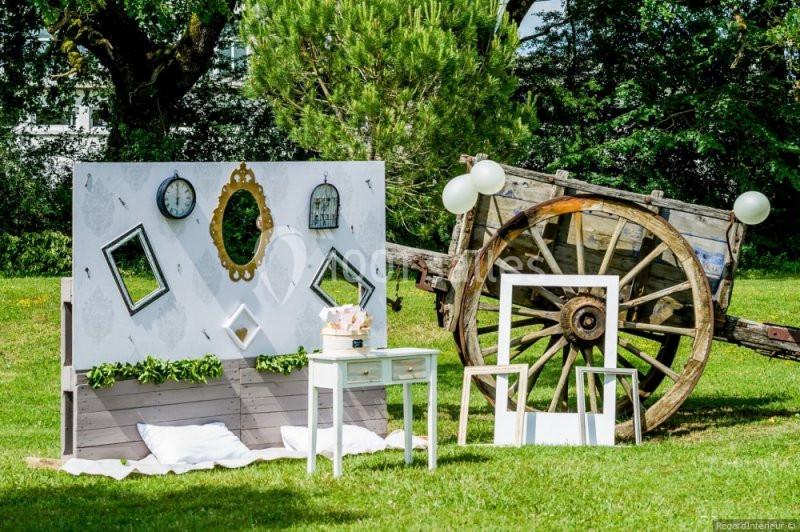Décor champêtre avec un panneau orné de miroirs et horloges, une table blanche et une charrette en bois sur pelouse.