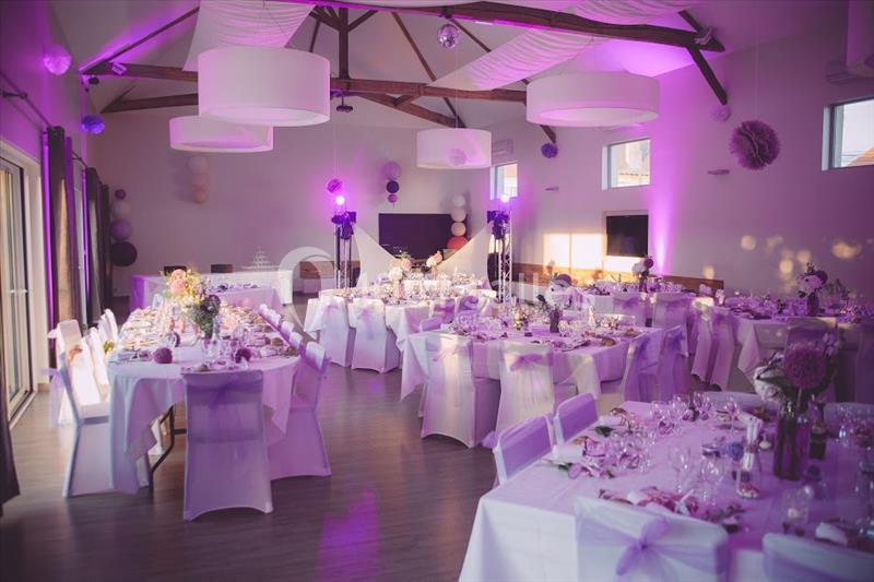 Salle de réception décorée en violet avec tables dressées, chaises ornées de rubans et éclairage tamisé.