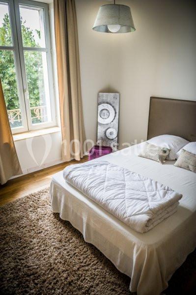 Chambre lumineuse avec lit double, couvre-lit plié, tapis beige, grande fenêtre et décoration murale.