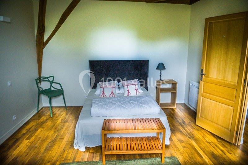 Chambre avec lit double, parquet en bois, table de chevet, lampe, chaise verte et banc au pied du lit.