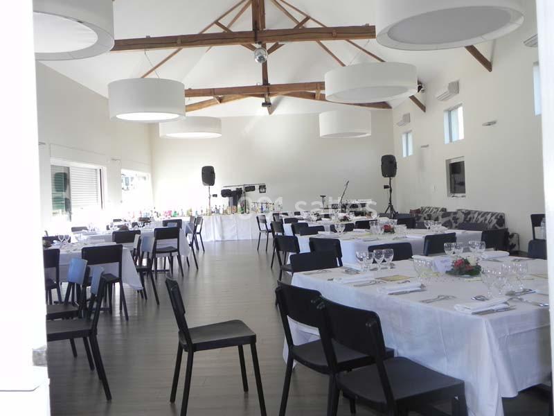 Salle de réception lumineuse avec tables dressées, chaises noires, poutres apparentes et espace pour un buffet.