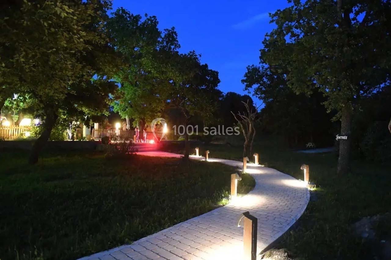 Chemin pavé éclairé par des lampes au sol, serpentant à travers un jardin arboré en soirée.