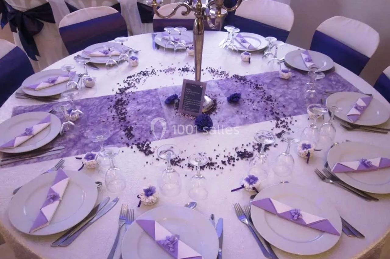Table ronde décorée pour un événement, avec nappe blanche, chemin de table violet, vaisselle et pliages de serviettes…