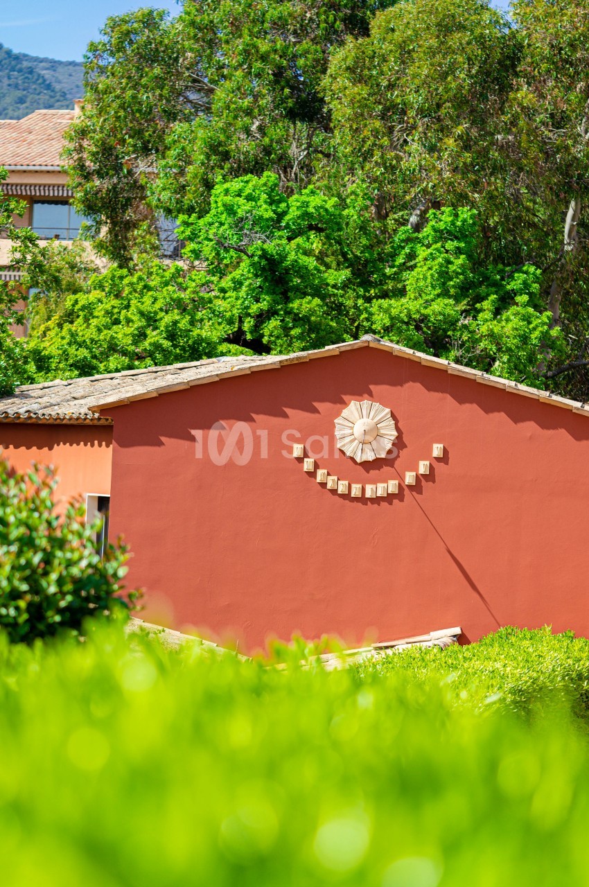 Façade d'une maison rouge ornée d'un décor en forme de sourire, entourée de verdure et d'arbres sous un ciel clair.