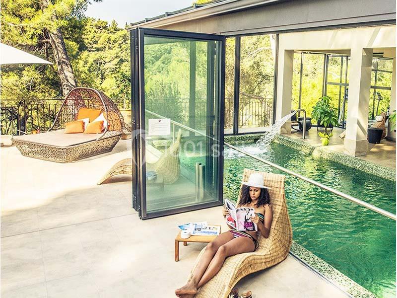Une femme lit sur une chaise longue près d'une piscine intérieure-extérieure entourée de végétation.