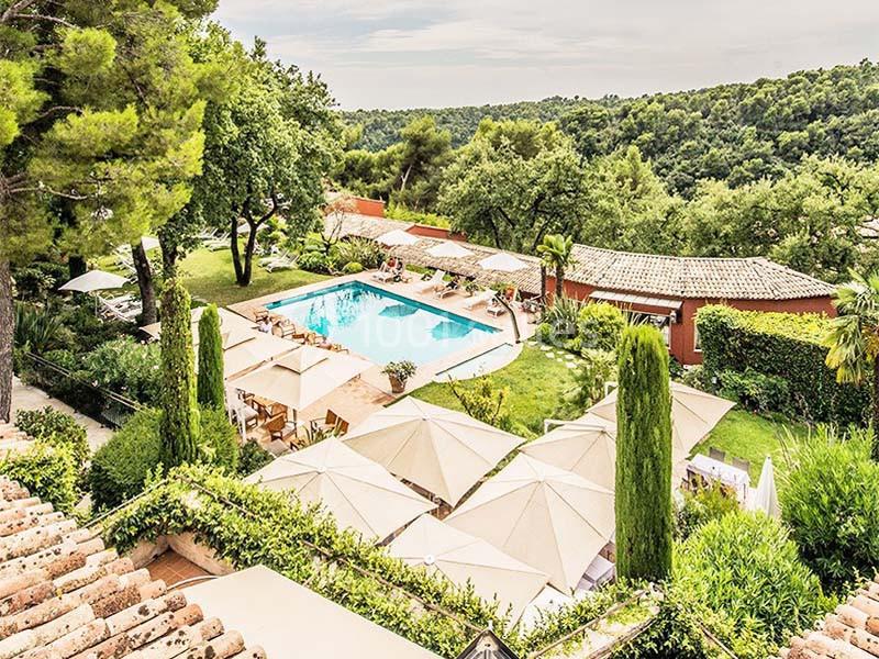 Vue d'une piscine entourée de verdure, de parasols et de bâtiments avec toits en tuiles dans un cadre naturel.