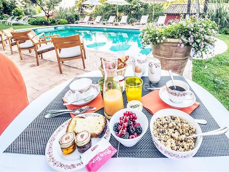 Petit-déjeuner servi en extérieur avec fruits, céréales et jus d'orange, près d'une piscine entourée de verdure.