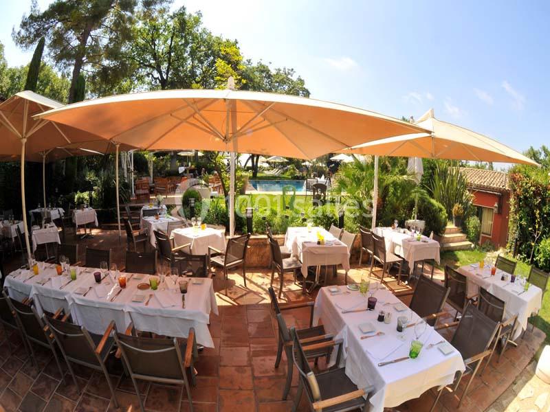 Terrasse d'un restaurant avec tables dressées sous de grands parasols, entourée de verdure et d'une piscine en arrière-plan.