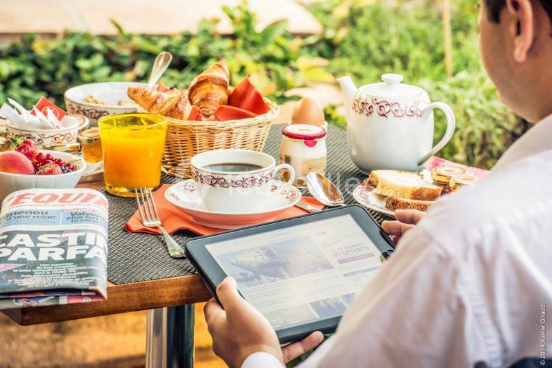Un homme lit sur une tablette à une table de petit-déjeuner avec croissants, café, jus d'orange et journal.
