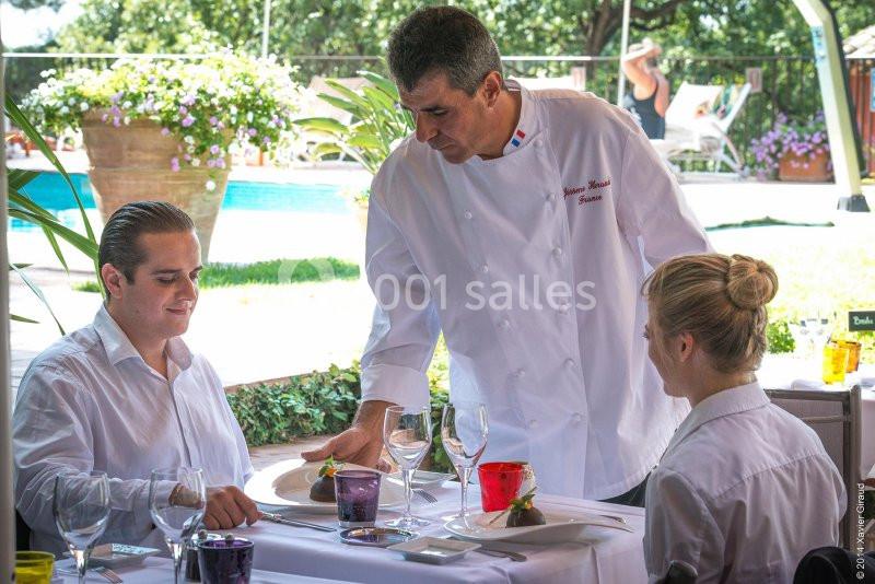 Un chef en tenue blanche sert un plat à un homme assis à une table en terrasse près d'une piscine.