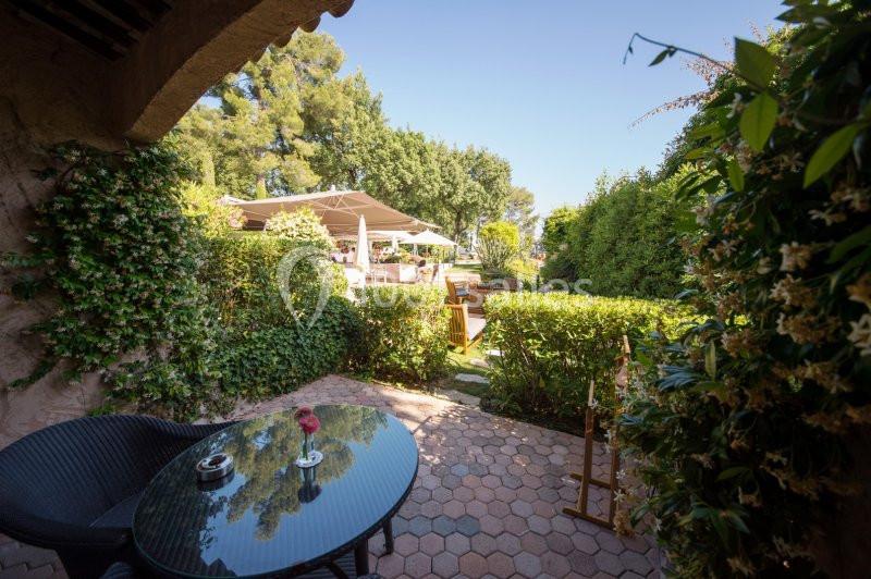 Terrasse ombragée avec table en rotin, vue sur un jardin verdoyant et des parasols au loin.