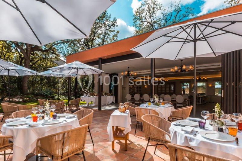 Terrasse d'un restaurant avec tables dressées, chaises en rotin et grands parasols blancs, entourée de verdure.