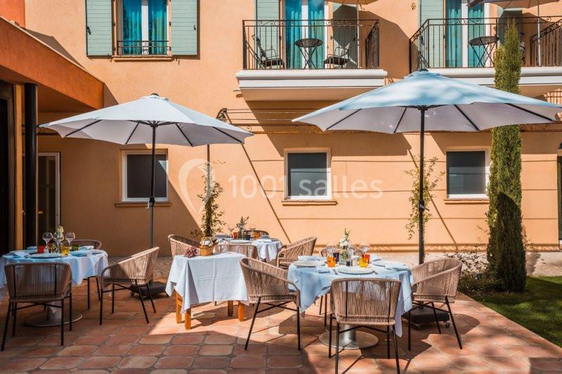 Terrasse d'un restaurant avec tables dressées, chaises en rotin et parasols blancs, devant une façade beige.