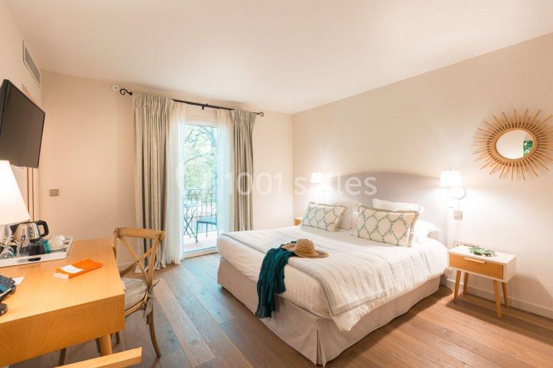 Chambre lumineuse avec lit double, bureau, télévision, balcon et décoration sobre dans des tons clairs.