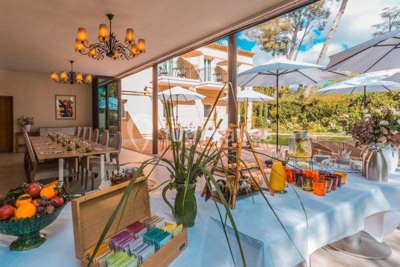 Table dressée avec fruits, boissons et infusions dans une salle lumineuse ouverte sur une terrasse avec parasols.
