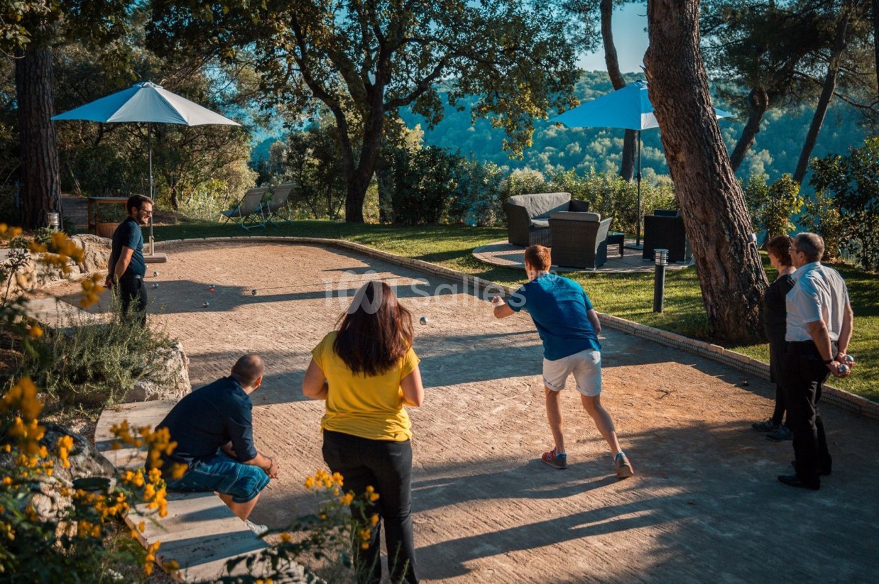 Des personnes jouent à la pétanque sur un terrain extérieur entouré de végétation et de parasols.