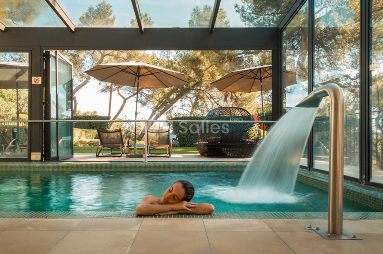 Femme se relaxant dans une piscine intérieure avec vue sur un jardin, sous une cascade d'eau.