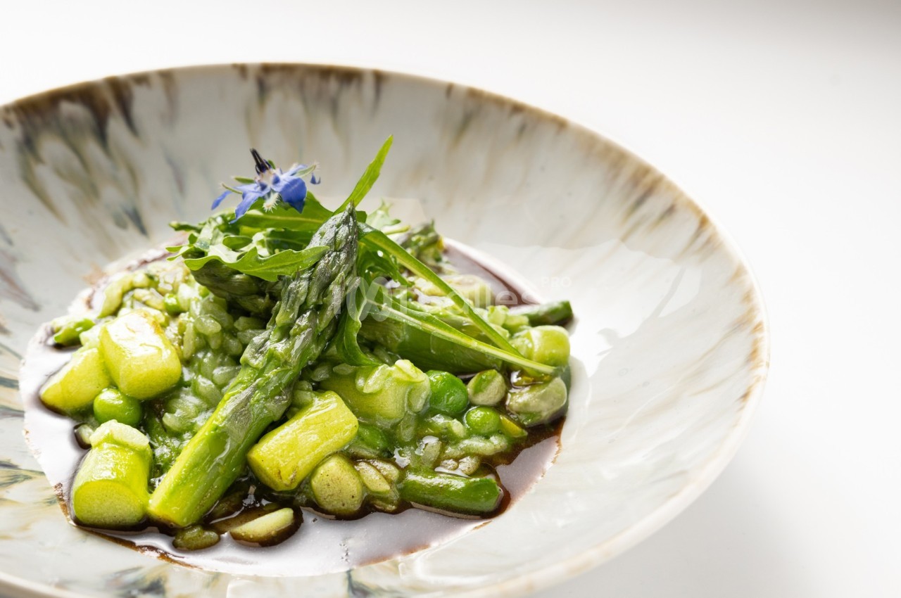 Assiette de risotto vert garni d'asperges, courgettes, graines et une fleur comestible, sur fond clair.