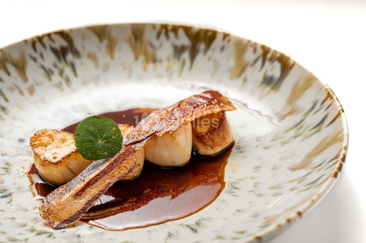 Noix de Saint-Jacques poêlées, accompagnées d'une sauce brune et d'une garniture croustillante dans une assiette décorative.