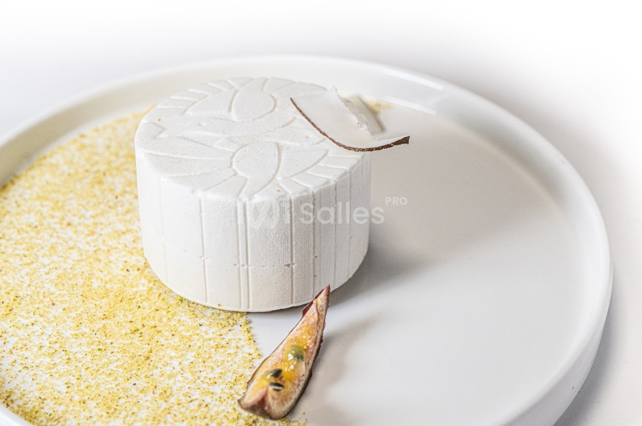 Dessert circulaire blanc texturé, présenté sur une assiette avec une poudre jaune et un morceau de fruit exotique.