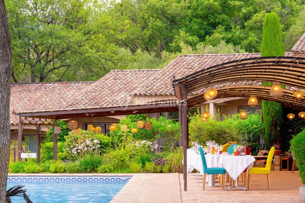 Terrasse extérieure avec tables dressées près d'une piscine, entourée de végétation et d'une pergola éclairée.