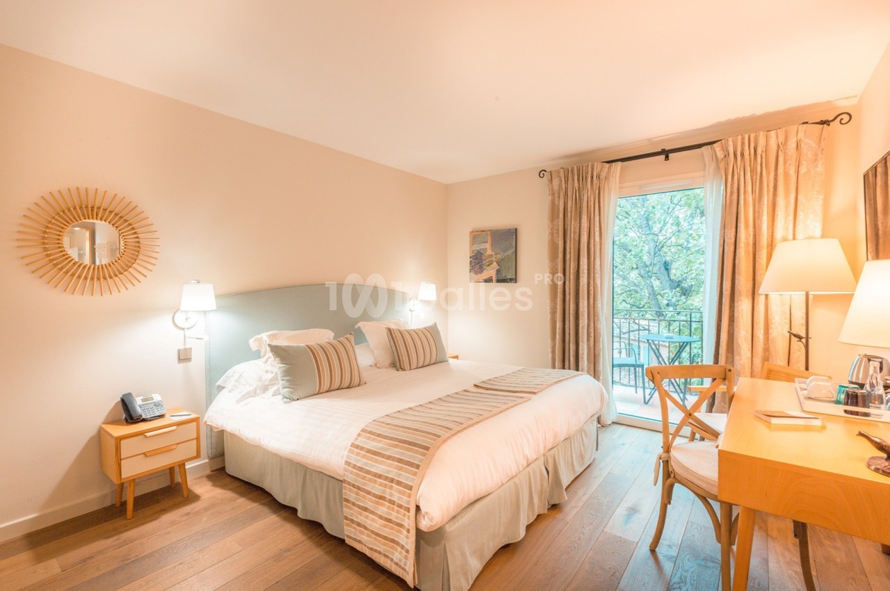 Chambre lumineuse avec lit double, bureau en bois, miroir rond et fenêtre donnant sur un balcon avec vue arborée.