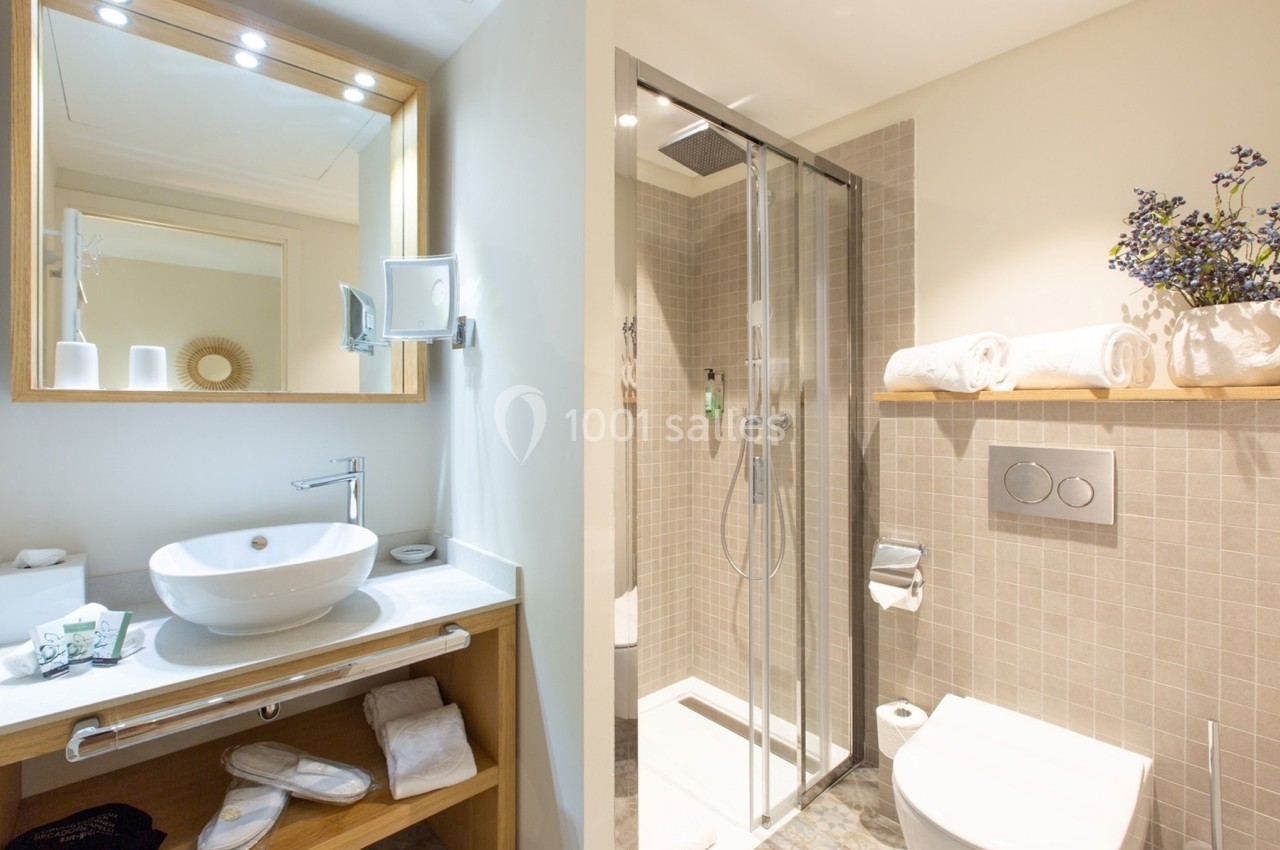 Salle de bain moderne avec douche vitrée, lavabo sur meuble en bois, miroir éclairé et étagère avec serviettes.