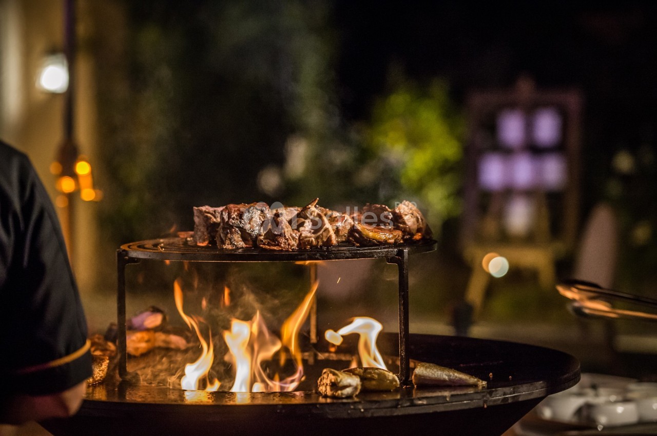 Viande grillant sur un barbecue au feu de bois, en extérieur, avec un arrière-plan flou et éclairé.