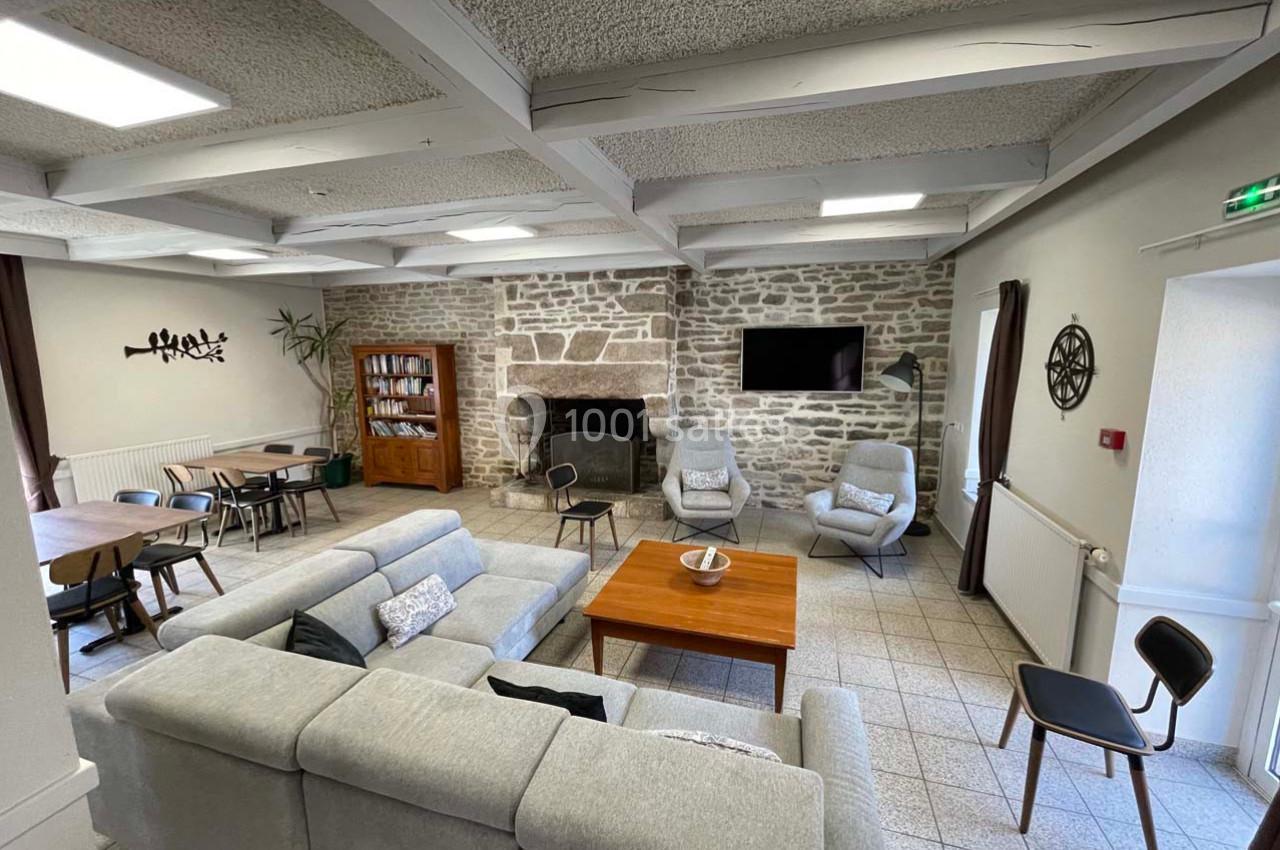 Salon spacieux avec canapé gris, table basse en bois, cheminée en pierre, télé murale et coin repas adjacent.