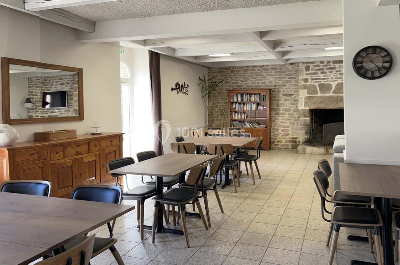 Salle lumineuse avec tables en bois, chaises noires, cheminée en pierre, bibliothèque et miroir sur un mur clair.