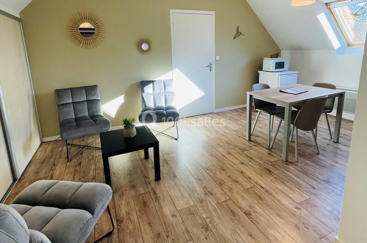 Salon lumineux avec parquet, fauteuils gris, table à manger, micro-ondes et décoration murale, sous une lumière naturelle.