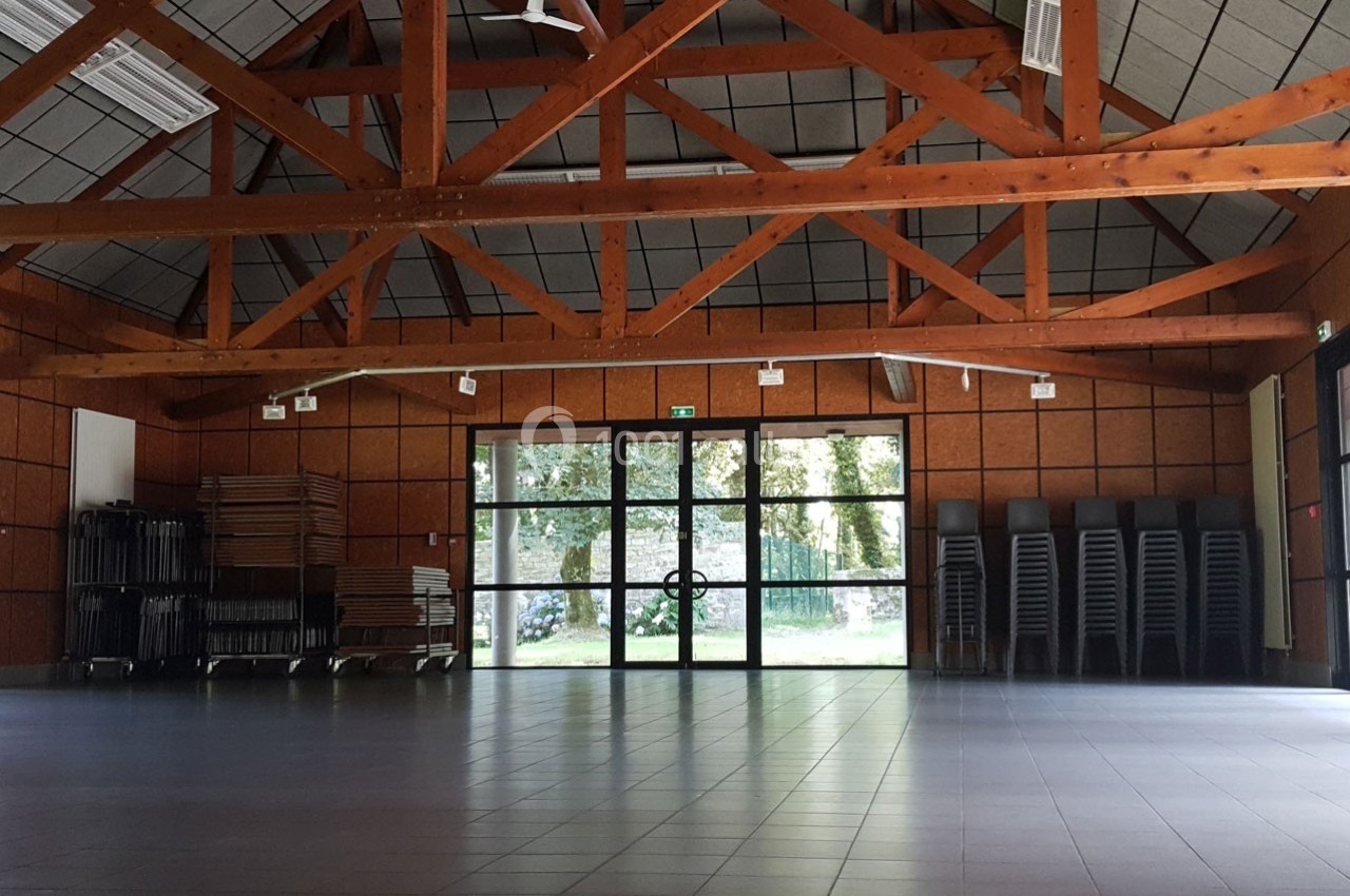 Salle vide avec charpente en bois, sol carrelé, chaises empilées et tables pliées près d'une baie vitrée donnant sur un…