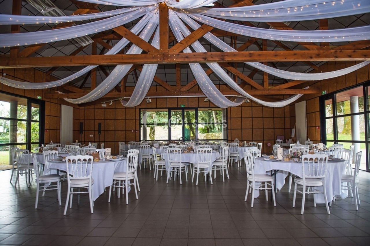 Salle de réception décorée avec des tables rondes dressées, des chaises blanches et des drapés suspendus au plafond.