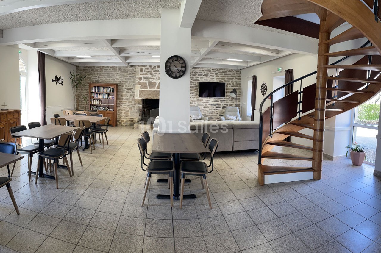 Salle lumineuse avec tables, chaises, canapé, escalier en bois, murs en pierre et grande horloge murale.
