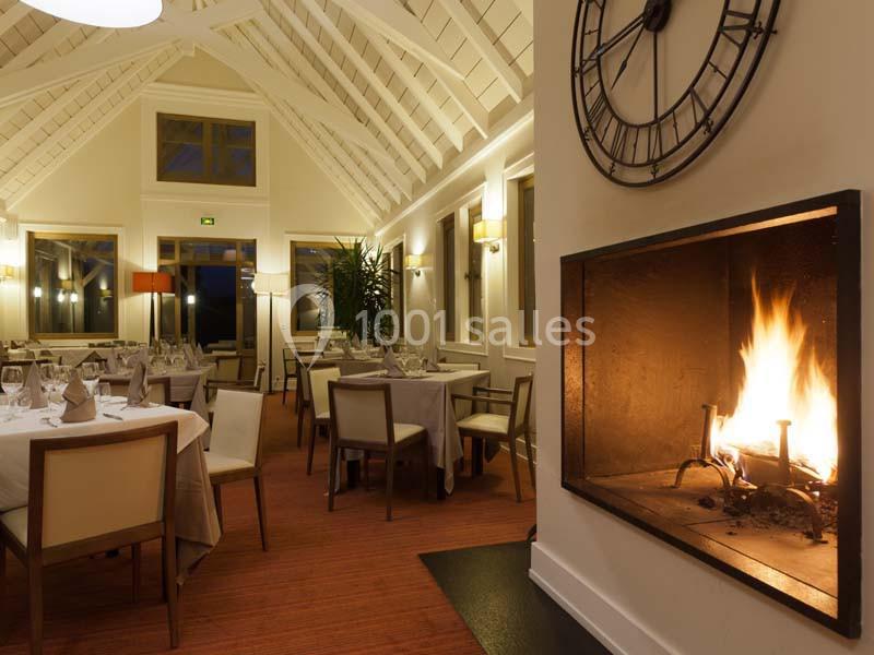 Salle de restaurant chaleureuse avec tables dressées, cheminée allumée et plafond voûté en bois blanc.