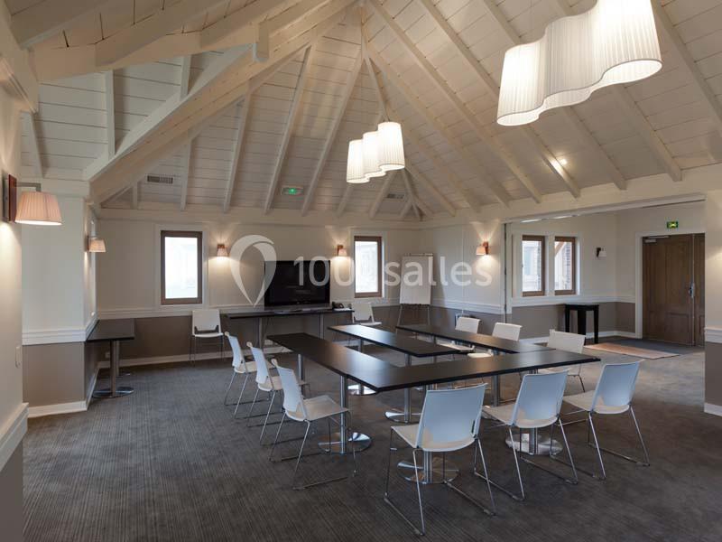 Salle de réunion lumineuse avec charpente apparente, tables noires, chaises blanches et éclairage suspendu.
