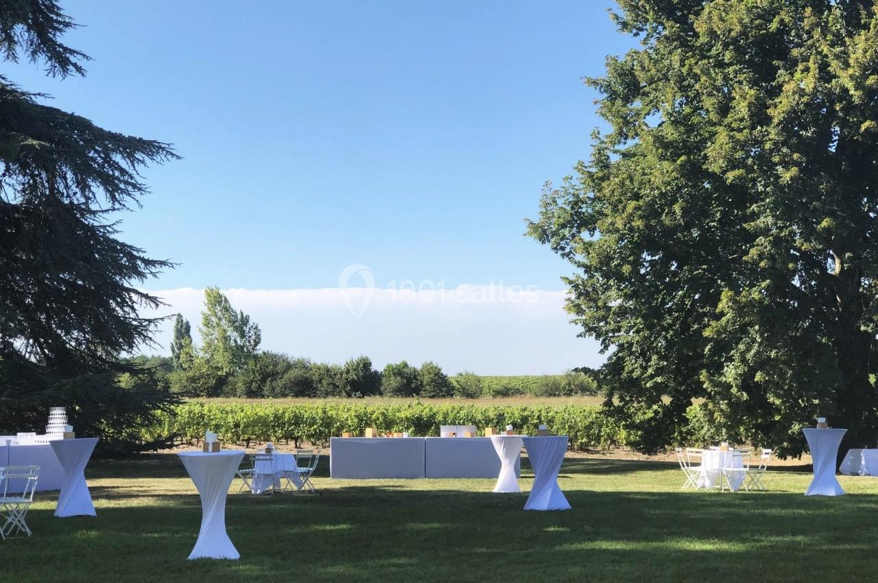 Tables hautes et chaises blanches disposées sur une pelouse, avec des arbres et des vignes en arrière-plan.