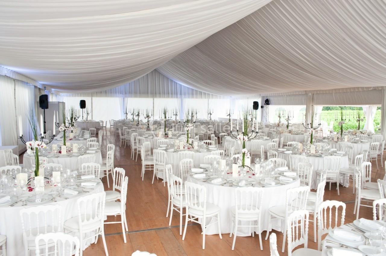Salle de réception sous une tente blanche avec tables rondes dressées, chaises blanches et décorations florales.