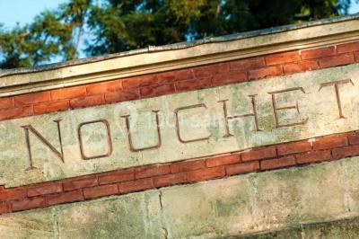 Inscription ’NOUCHET’ gravée sur une façade en pierre et briques, entourée de végétation en arrière-plan.