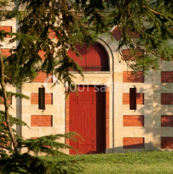 Façade en briques et pierre avec une porte rouge, entourée de verdure et partiellement ombragée par des branches.