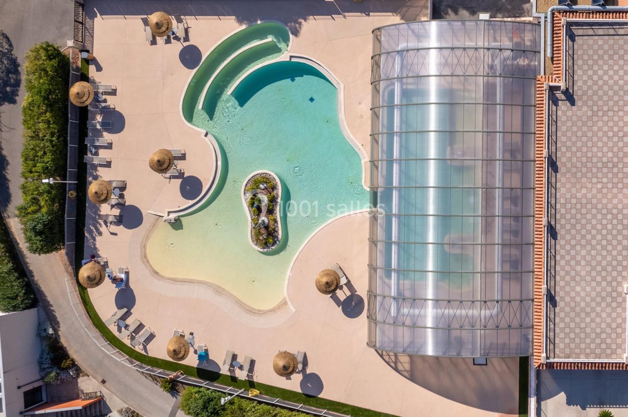 Vue aérienne d'une piscine extérieure avec une partie couverte, entourée de parasols et de chaises longues.
