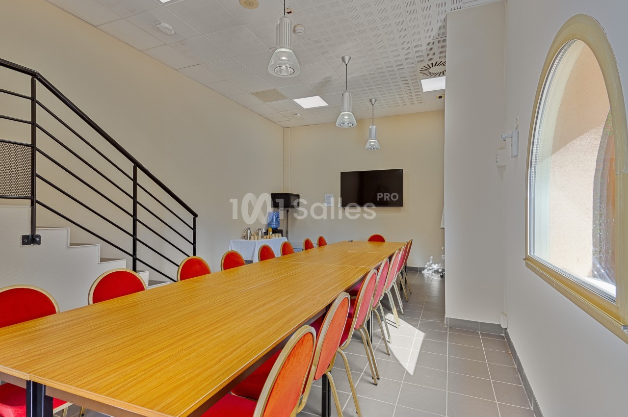 Salle de réunion lumineuse avec une grande table en bois, des chaises rouges, un écran mural et un coin boissons.