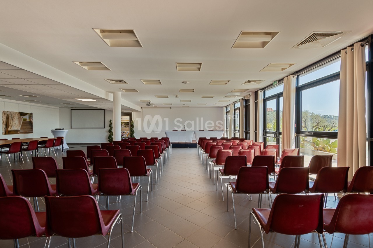 Salle de conférence lumineuse avec des rangées de chaises rouges, un tableau blanc et de grandes fenêtres.