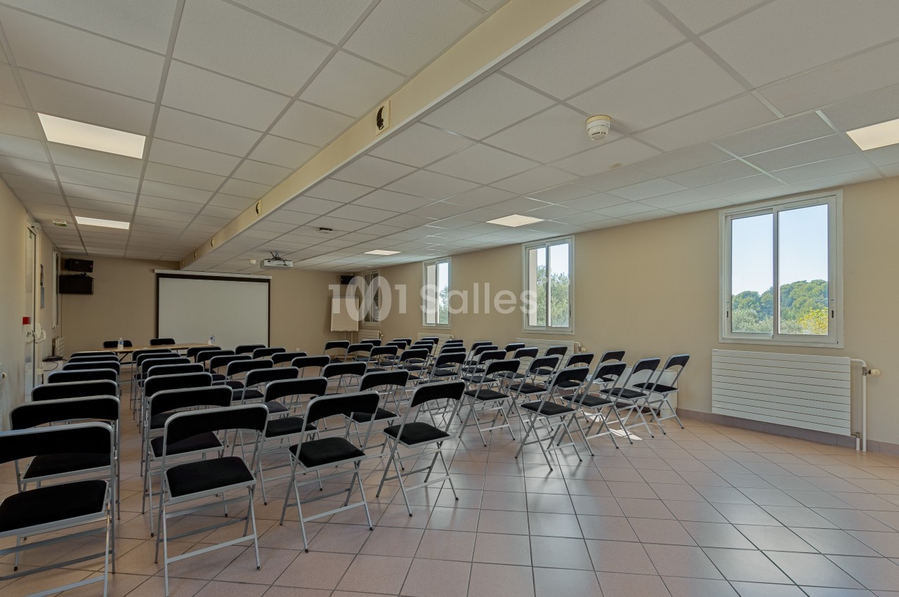 Salle de réunion lumineuse avec des rangées de chaises pliantes face à un écran de projection.