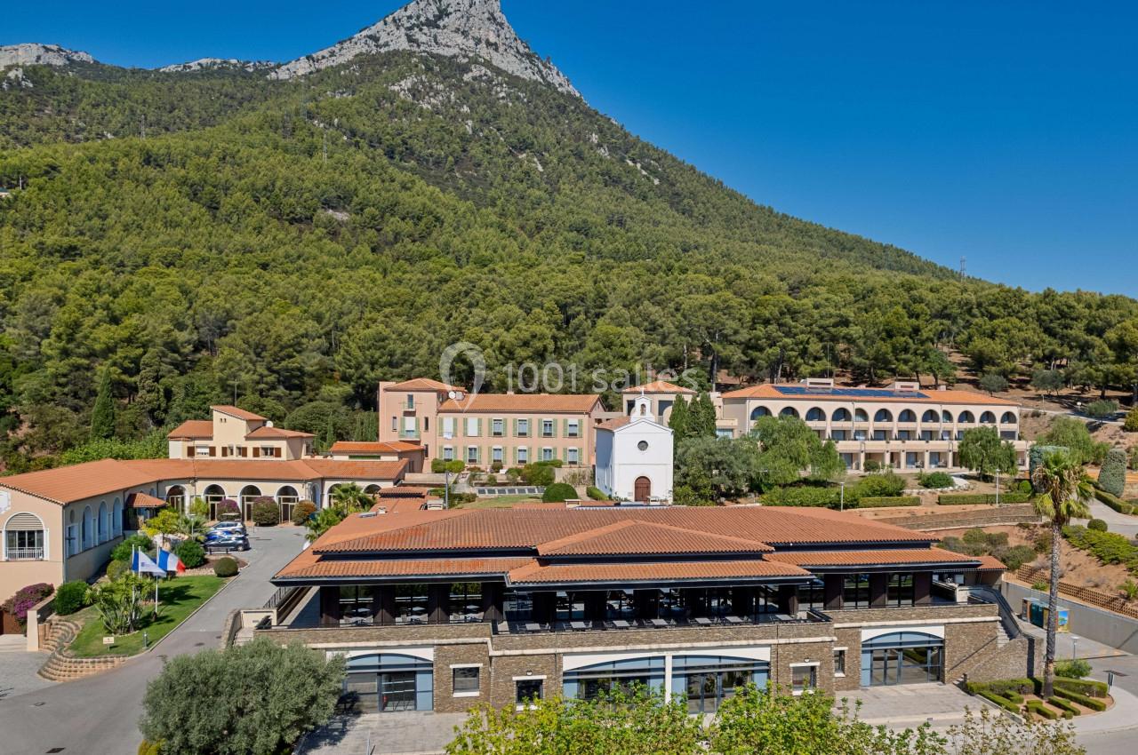 Vue aérienne d'un complexe de bâtiments entouré de végétation, avec une montagne en arrière-plan sous un ciel dégagé.