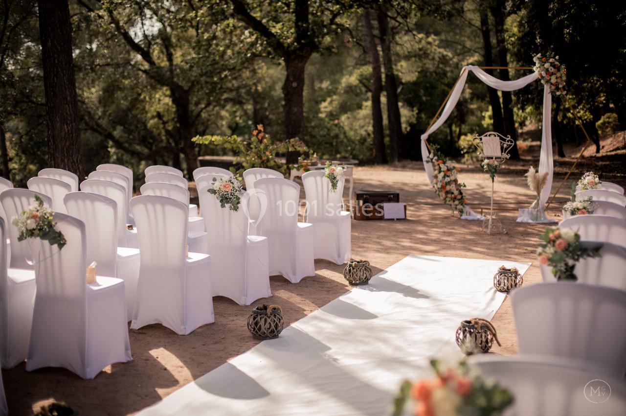 Chaises blanches alignées en extérieur, décorées de fleurs, face à une arche ornée pour une cérémonie en plein air.